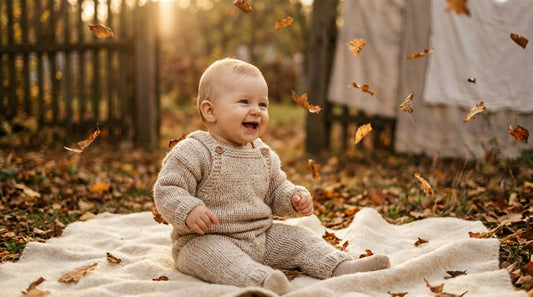 Baby im naturfarbenen Strick-Overall sitzt lachend auf einer Wolldecke im herbstlichen Garten